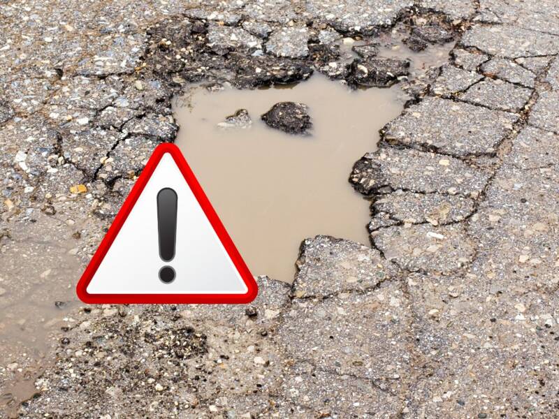 Ein Schlagloch in einer asphaltierten Straße, gefüllt mit Wasser, ist mit einem Warnsymbol markiert.