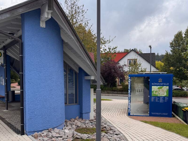 Ein blaues, überdachtes Bushäuschen mit einer Landkarte und Sitzmöglichkeiten steht an einem gepflasterten Weg, umgeben von Bäumen und Pflanzen.