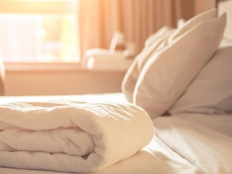 Ein frisch gefaltetes weißes Handtuch liegt auf einem ordentlich gemachten Bett in einem von Sonnenlicht durchfluteten Raum.