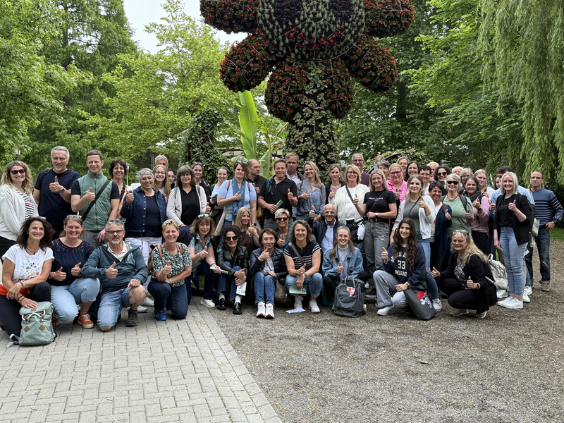 Eine Gruppe von über vierzig lächelnden Menschen posiert in Freizeitkleidung vor einer großen Blumenstruktur in einem grünen Park und zeigt Daumen hoch.