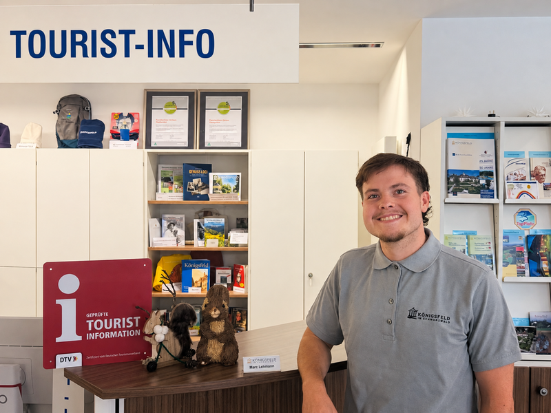Ein lächelnder Mitarbeiter steht an einem Empfangstresen in einer Tourist-Info, umgeben von Broschüren und Souvenirs.