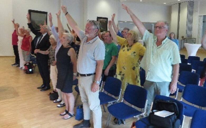Eine Gruppe von Menschen steht in einem Raum und hält die rechte Hand mit gestrecktem Arm nach oben.