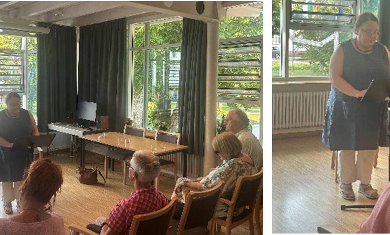 Eine Person hält eine Präsentation oder Lesung vor einem kleinen Publikum in einem Raum mit großen Fenstern.