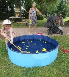 Ein Kind spielt mit einer Angelrute an einem blauen Planschbecken voller gelber Spielenten im Freien, während im Hintergrund eine Frau und eine weitere Person mit einem Kinderwagen stehen.