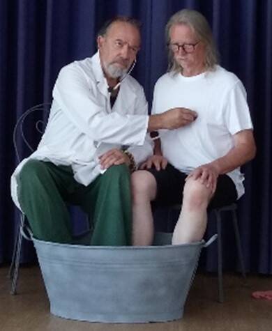 Zwei Männer sitzen in einem Raum, wobei der linke Mann in einem weißen Kittel den Puls des rechten Mannes in einem weißen T-Shirt misst, während dieser seine Füße in einer großen Metallwanne mit Wasser hat.