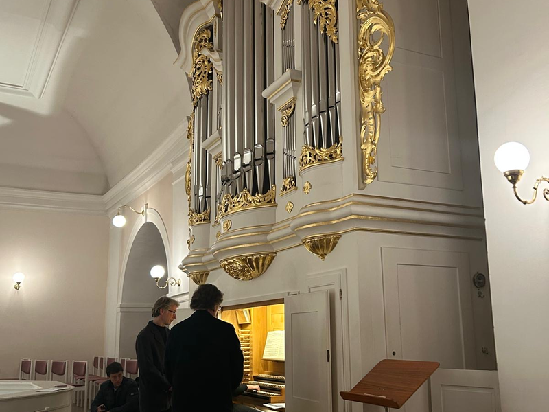 Ein prachtvolles Orgelgehäuse mit goldenen Verzierungen befindet sich oberhalb eines Raumes, in dem mehrere Personen stehen.