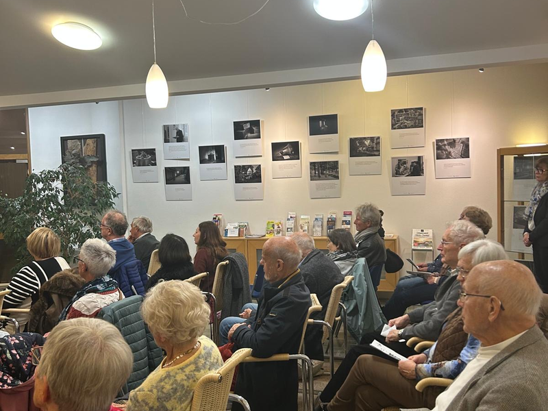 Eine Gruppe von Menschen sitzt in einem Raum und schaut nach vorne, während an der Wand Bilder hängen.
