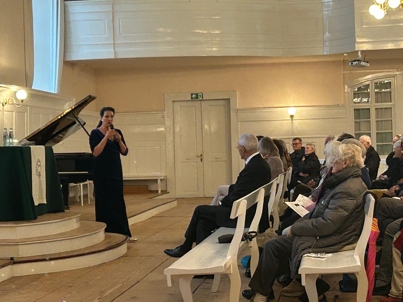 Eine Frau im langen, schwarzen Kleid spricht in einem Saal vor einem sitzenden Publikum, während ein Konzertflügel auf der Bühne steht.