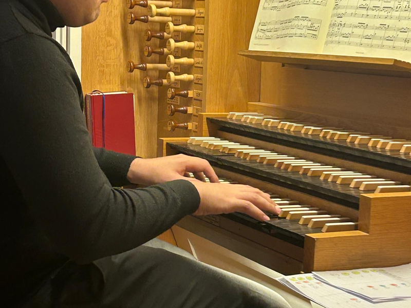 Eine Person spielt konzentriert an einem Orgelspieltisch, umgeben von Notenblättern und Orgelzügen.