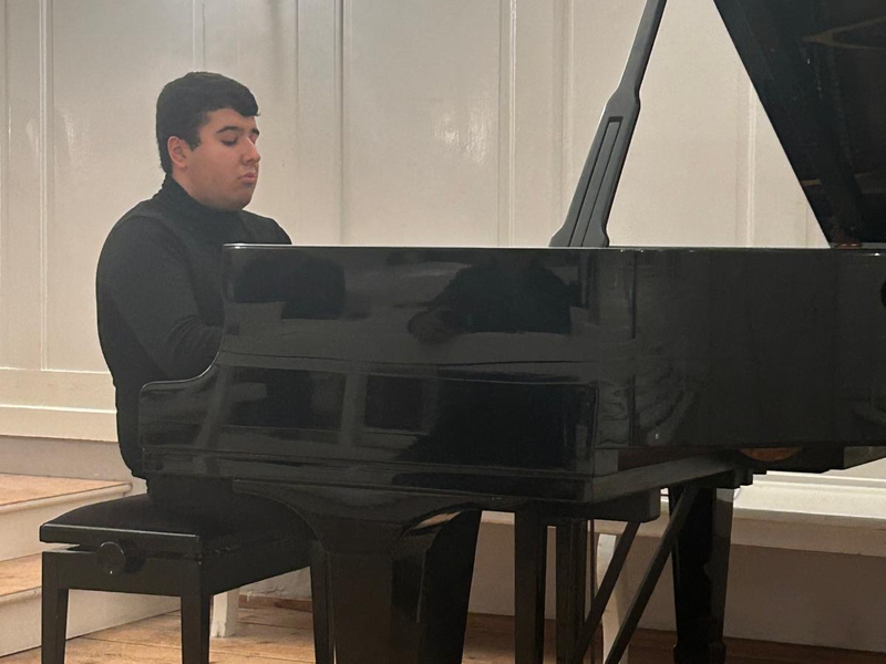 Eine Person spielt Klavier auf einer Bühne in einem Raum mit weißer Wandverkleidung.