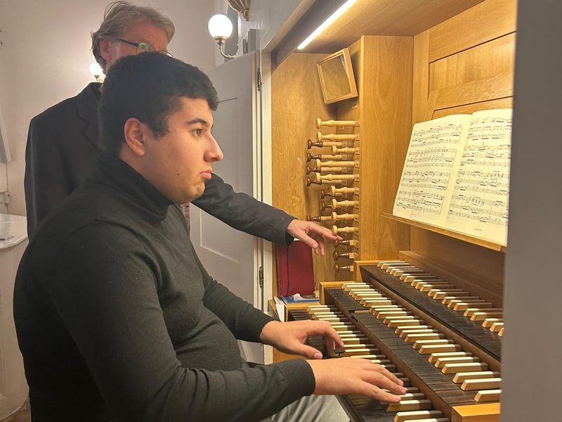 Eine Person spielt Orgel in einem Innenraum, während eine andere daneben steht und zuschaut.