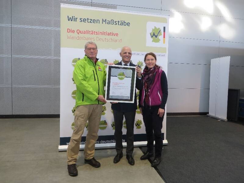Drei Personen stehen vor einem Banner mit der Aufschrift "Wanderbares Deutschland" und halten eine Auszeichnung.