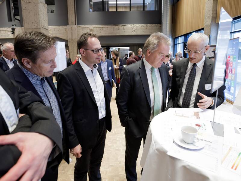 Eine Gruppe von Männern in Anzügen steht zusammen und betrachtet ein Dokument auf einem Tisch bei einer Veranstaltung in einem modern gestalteten Raum.