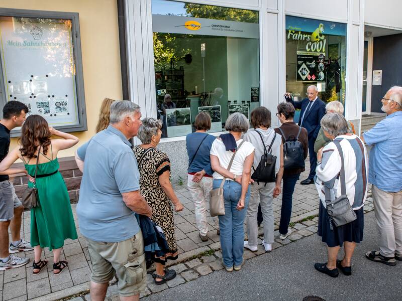 Eine Gruppe von Menschen steht vor einem Schaufenster und hört einer Person zu, die drinnen zu ihnen spricht.