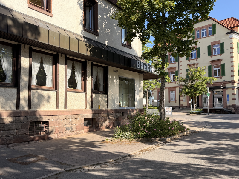 Das Bild zeigt eine Straßenecke mit einem Gebäude, auf dem ein Schild mit der Aufschrift "Bäckerei" hängt, umgeben von einigen Bäumen und weiteren mehrstöckigen Wohnhäusern im Hintergrund.