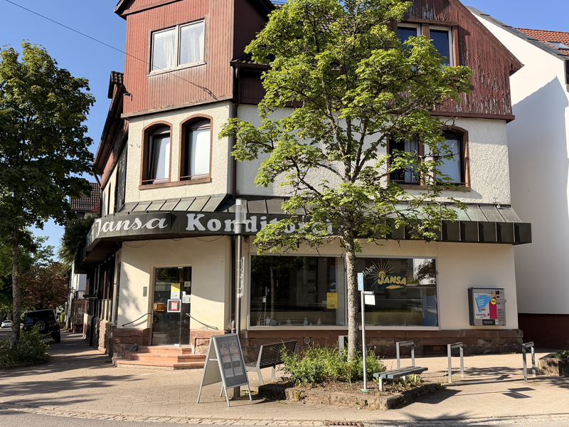 Ein zweistöckiges Gebäude an einer Straßenecke, das mit dem Schriftzug "Jansa Konditorei" versehen ist, steht hinter einem Baum und einem Gehweg.