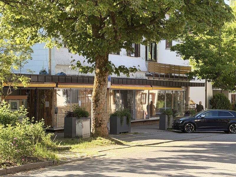 Ein älteres Gebäude mit großen Glasfronten und einem geparkten schwarzen Auto davor, umgeben von grünen Bäumen.