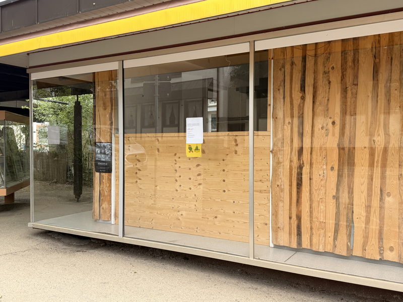 Ein moderner Holzpavillon mit großen Glastüren und einer Holzwand dahinter, im Vordergrund ein Schaukasten.