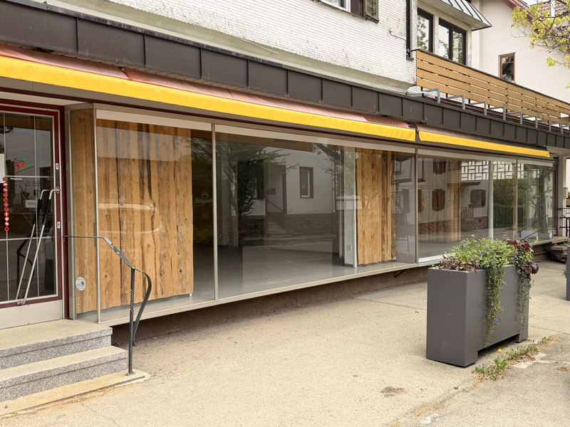 Ein leerstehendes Geschäft mit großen Glasfenstern und einer Holzwand, flankiert von einem Pflanzkübel.