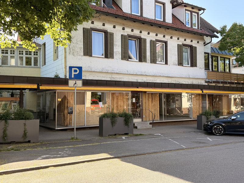 Ein zweistöckiges Gebäude mit weißen Wänden und braunen Fensterläden hat im Erdgeschoss eine glasverkleidete Fassade und davor ist ein Parkplatz mit einem Behindertenparkplatzschild.
