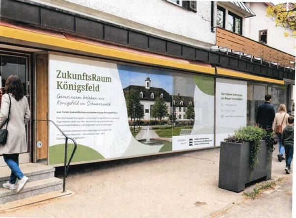 Eine Frau betritt ein Gebäude mit einem großen Werbeplakat für den "ZukunftsRaum Königsfeld", das eine Fotografie eines stattlichen Gebäudes und Text über ein Gemeinschaftsprojekt zeigt.