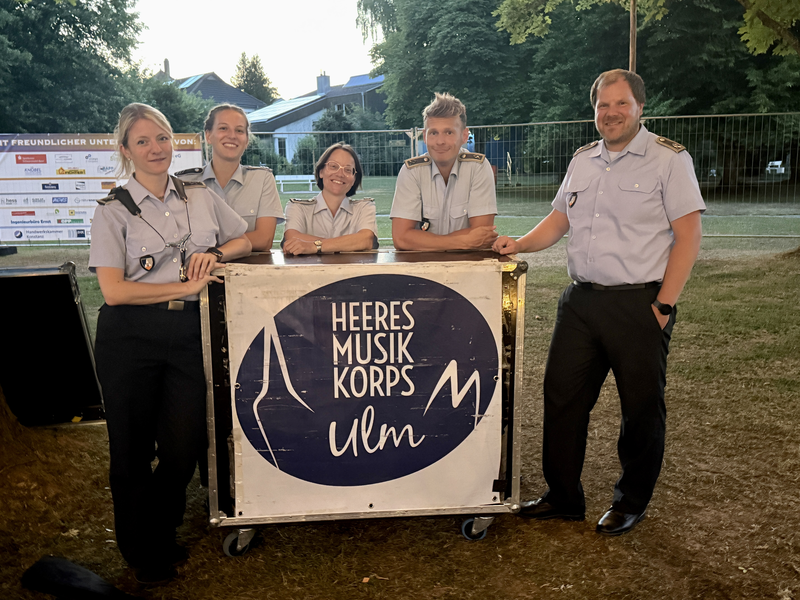 Fünf Personen in Uniform posieren lächelnd um einen mit "Heeresmusikkorps Ulm" beschrifteten Kasten in einem Freienbereich.