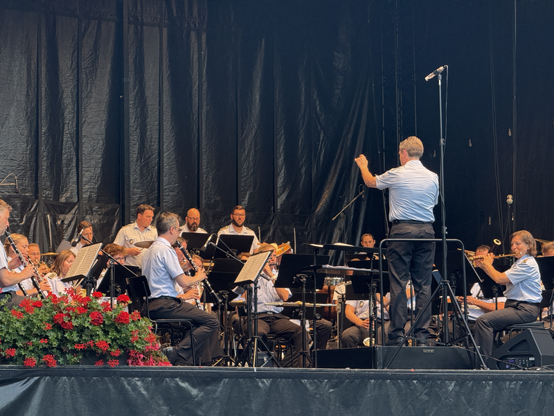 Ein Orchester spielt auf einer Bühne, geleitet von einem Dirigenten, umgeben von Blumenarrangements.