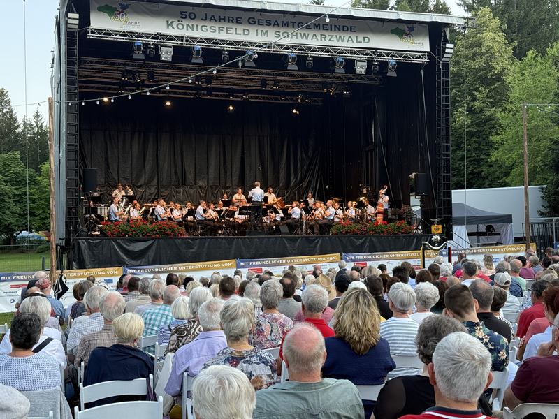 Eine große Menschenmenge sitzt vor einer Bühne im Freien, auf der ein Orchester mit einem Dirigenten spielt.