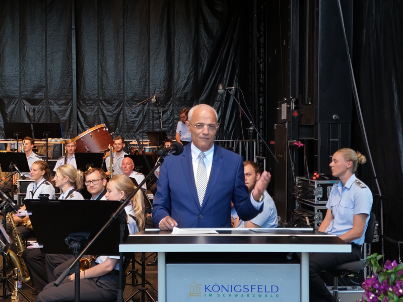 Ein Mann in einem blauen Anzug spricht an einem Pult vor einem Orchester auf einer Bühne.