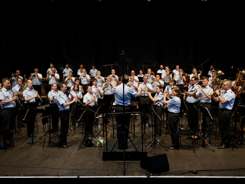 Eine große Blaskapelle, bestehend aus Musikern in einheitlicher Kleidung, spielt auf einer Bühne unter der Leitung eines Dirigenten.