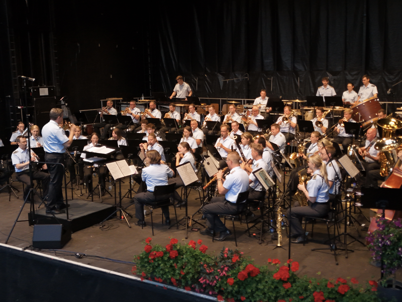Eine große Blaskapelle, bestehend aus Musikern in einheitlicher Kleidung, spielt auf einer Bühne unter der Leitung eines Dirigenten.