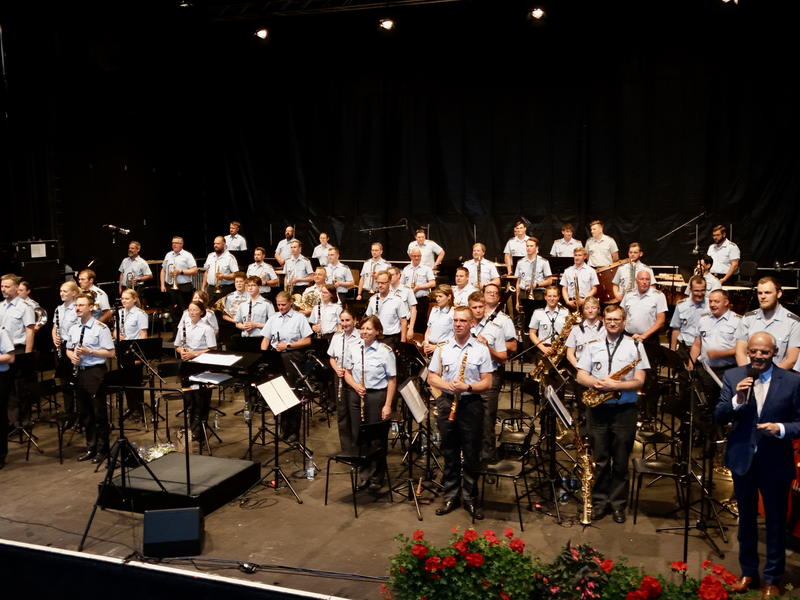 Eine große Blaskapelle, bestehend aus Musikern in einheitlicher Kleidung, spielt auf einer Bühne unter der Leitung eines Dirigenten.