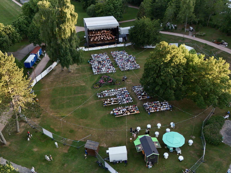 Eine Open-Air-Veranstaltung mit einer Bühne und vielen Besuchern, die auf Stühlen auf einer großen, von Bäumen umgebenen Wiese sitzen.