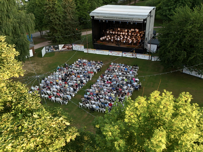 Eine Open-Air-Veranstaltung mit einer Bühne und vielen Besuchern, die auf Stühlen auf einer großen, von Bäumen umgebenen Wiese sitzen.