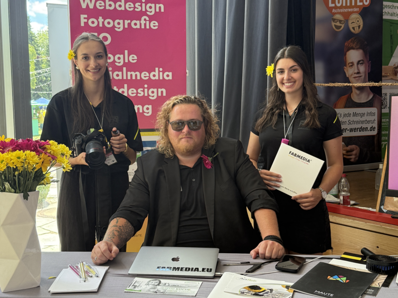 Drei Personen stehen hinter einem Tisch mit Informationsmaterialien und einem Laptop, umgeben von bunten Blumen und Werbebannern, vermutlich auf einer Messe oder Veranstaltung.