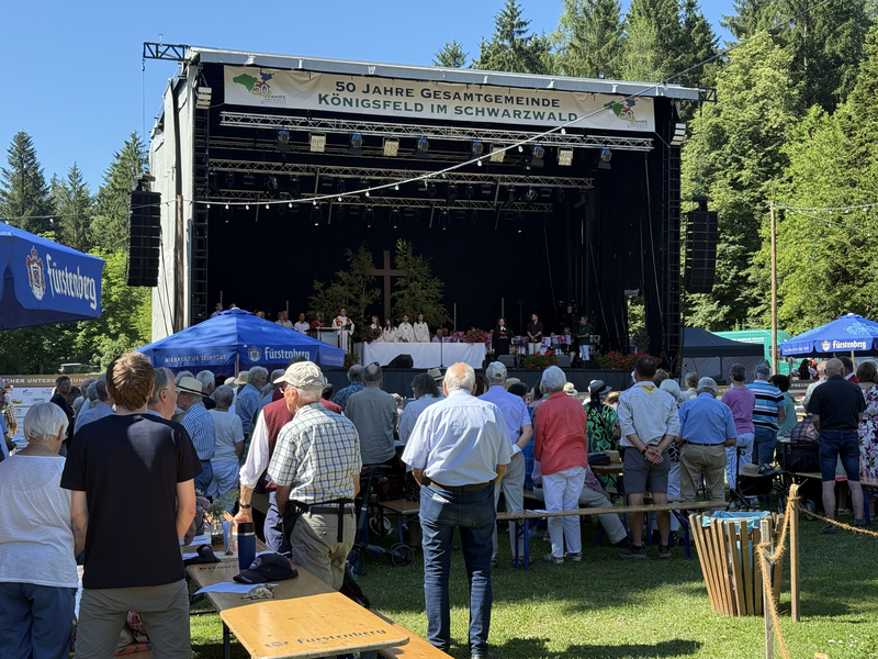 Eine große Menschenmenge versammelt sich vor einer Bühne im Freien für eine Feier im Grünen, mit einem Banner, das "50 Jahre Gesamtgemeinde Königsfeld im Schwarzwald" verkündet.