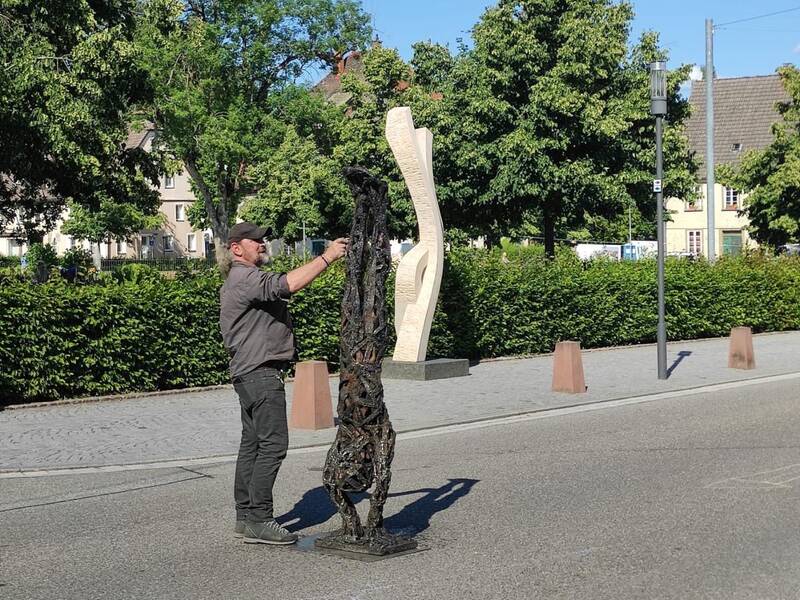 Ein Mann in Freizeitkleidung steht in einer gepflasterten städtischen Umgebung und arbeitet an einer skulpturalen Struktur, während im Hintergrund Bäume und eine weitere abstrakte Skulptur zu sehen sind.