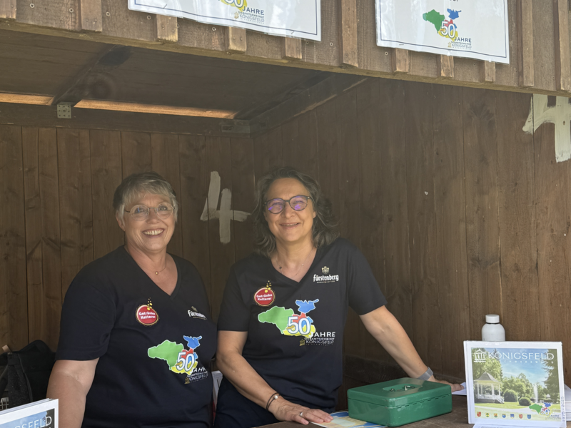 Zwei lächelnde Personen stehen hinter einem Holztresen mit Flyern und Broschüren, über dem ein Schild mit der Aufschrift "Jubiläums-Chronik 35,00 €" hängt.