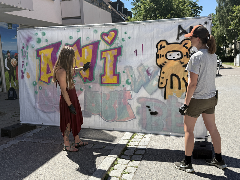 Zwei Personen betrachten eine bemalte Leinwand im Freien, die bunte Graffiti und eine Zeichnung eines bärähnlichen Wesens zeigt.