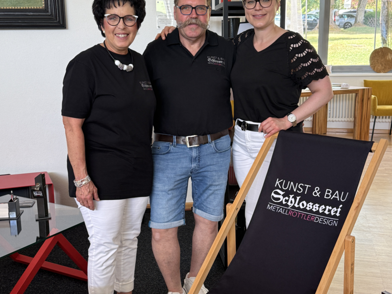 Drei Personen stehen nebeneinander in einem Innenraum, tragen schwarze Shirts mit einem Firmenlogo und lächeln in die Kamera; im Vordergrund ist ein Liegestuhl mit der Aufschrift "Kunst & Bau Schlosserei Metall Rottler Design" zu sehen.