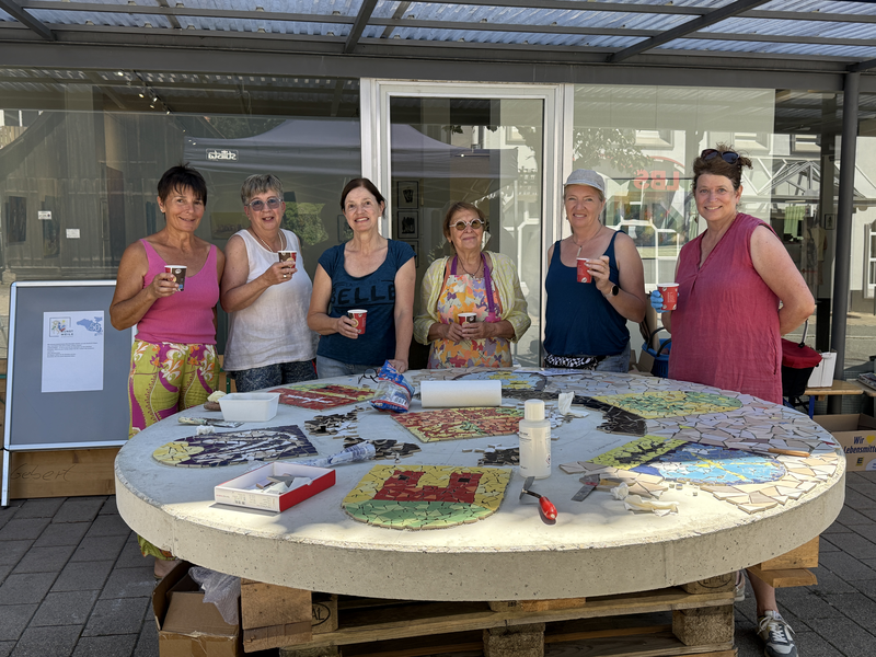 Sechs Menschen stehen lächelnd um einen runden Tisch mit einem Mosaik-Projekt und halten Pappbecher in der Hand.