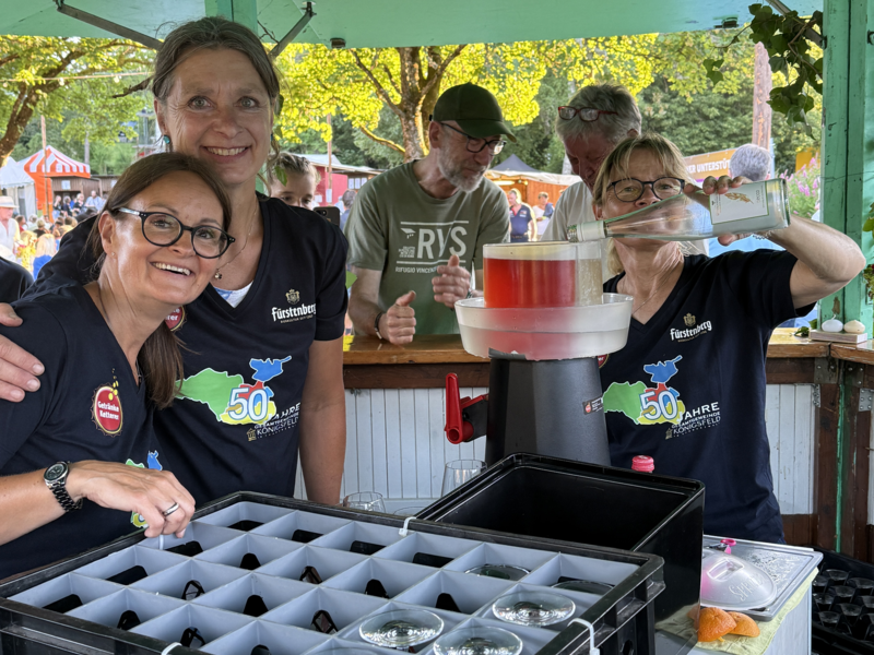 Drei Personen stehen in einem Getränkestand bei einem Fest und lächeln, während eine von ihnen ein Getränk in ein Glas einschenkt.
