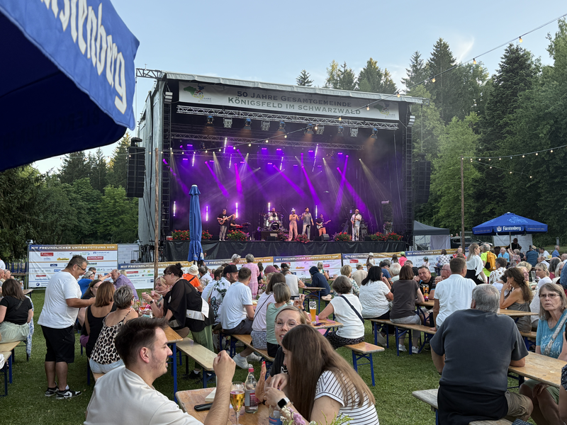 Auf einer Bühne spielt eine Band vor einem Publikum, das auf Bierbänken im Freien sitzt und die Veranstaltung genießt.