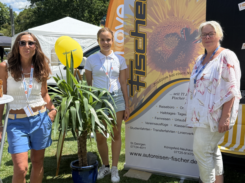 Drei Personen stehen auf einer Wiese neben einem Werbebanner mit Sonnenblumenmotiv, einem gelben Ballon und einer Pflanze, während im Hintergrund Bäume und ein Zelt zu sehen sind.