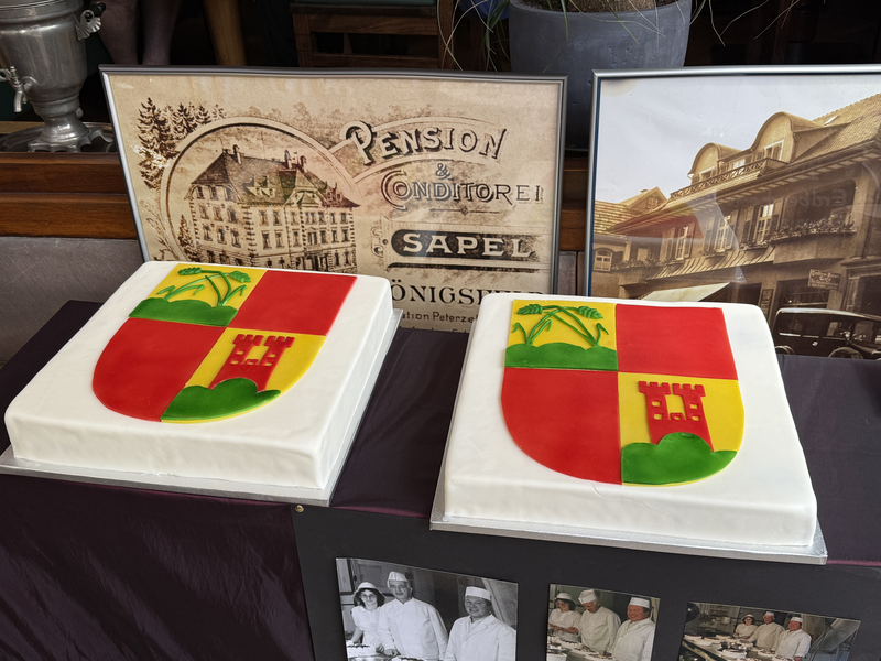 Zwei rechteckige Torten mit einem farbigen Wappen darauf stehen vor nostalgischen Bildern einer Konditorei.