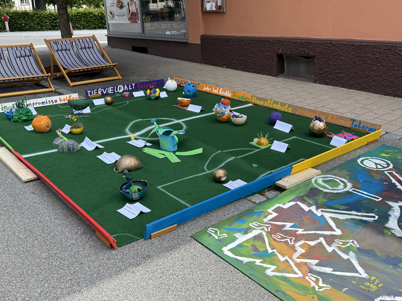 Ein kunstvoll gestaltetes Miniatur-Fußballfeld ist auf dem Boden mit verschiedenen bunten Objekten, darunter Gießkannen und Kugeln, dekoriert und von bemalten Holzlatten umrahmt; im Hintergrund sind zwei Liegestühle zu sehen.