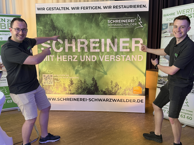 Zwei Personen stehen lächelnd vor einem Werbebanner der Schreinerei Schwarzwälder, das mit einem Waldmotiv und dem Schriftzug "Schreiner mit Herz und Verstand" bedruckt ist.
