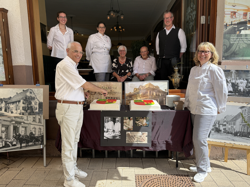 Eine Gruppe von Menschen steht lächelnd vor einem Tisch mit einer bunt dekorierten Torte und alten Fotografien, die an eine historische Feier erinnern.