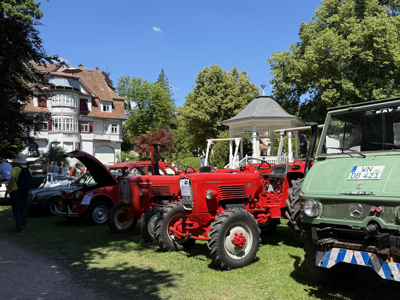 Mehrere Vintage-Fahrzeuge, darunter rote Traktoren und ein grünes Fahrzeug, sind auf einer Grünfläche vor einem großen Haus und einem Pavillon ausgestellt.