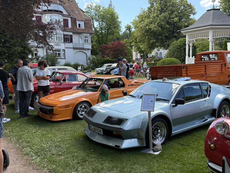 Mehrere Menschen betrachten eine Ausstellung von Oldtimern auf einem grünen Rasen vor einem eleganten, älteren Gebäude und einem Pavillon.